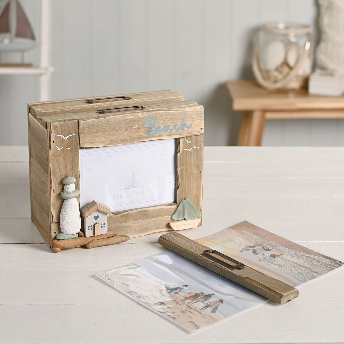 Driftwood photo trinket box with rectangular window, Beach text, pebble lighthouse, wooden house, and sea glass boat detail, shown open on a table with photo cards nearby.