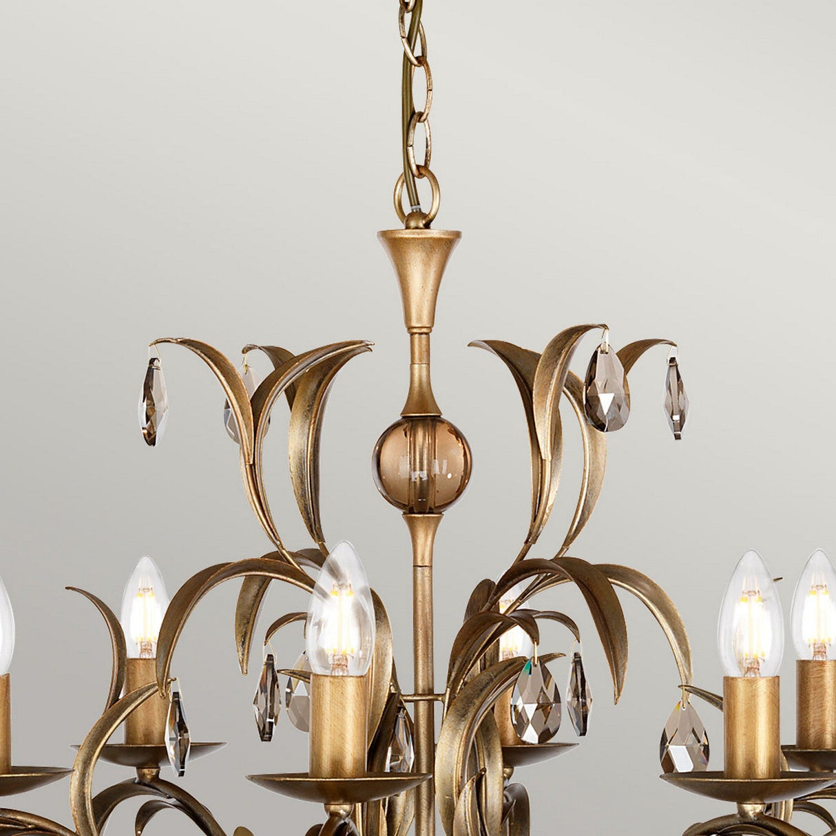A close-up of the Lily 8 Light Chandelier - Bronze Patina highlights its intricate bronze patina finish with gracefully curved arms embellished with smoked cut glass droplets. The chandelier features multiple candle-style light bulbs, providing a vintage and luxurious look against a light gray background.