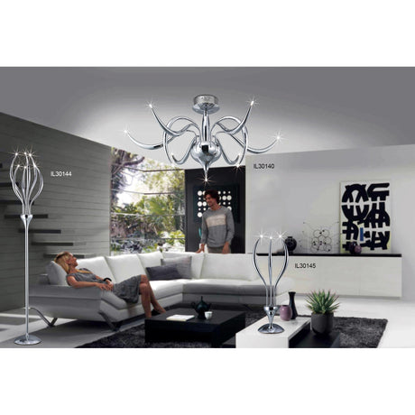 A modern living room with gray and white furniture, abstract wall art, large windows, and the Llamas 3 Light Table Lamp in polished chrome. A woman relaxes on a couch while another stands nearby.