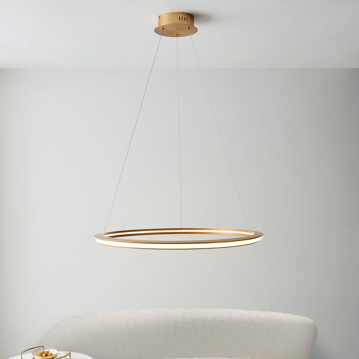 Lighting No.1 Ashenhurst LED ring pendant in satin gold, suspended from a ceiling plate by thin cables, hanging above a pale upholstered chair.