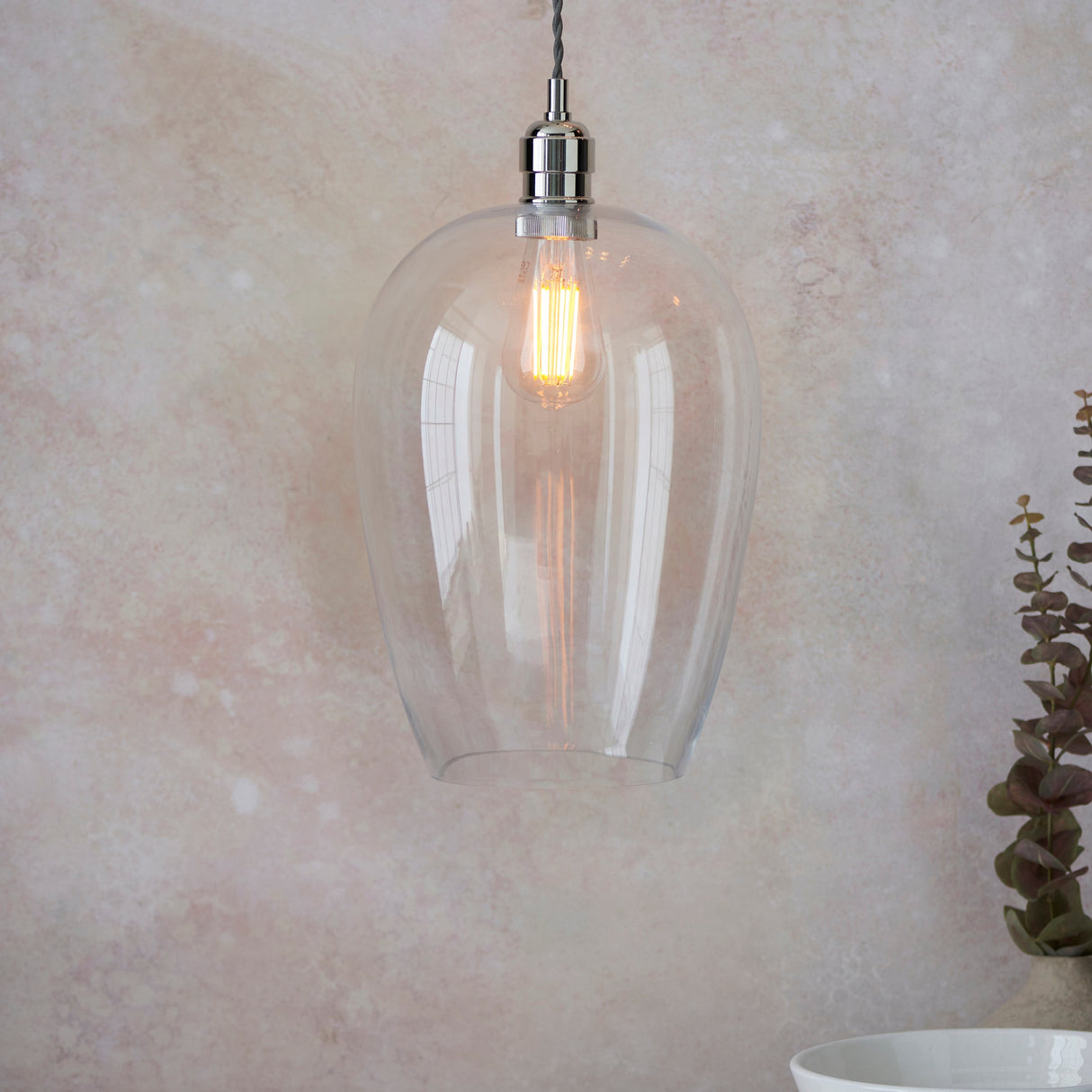Vellmere Large Pendant by Lighting No.1 with clear glass shade and bright nickel fitting, lit filament bulb, hung against a pink-toned wall with eucalyptus.