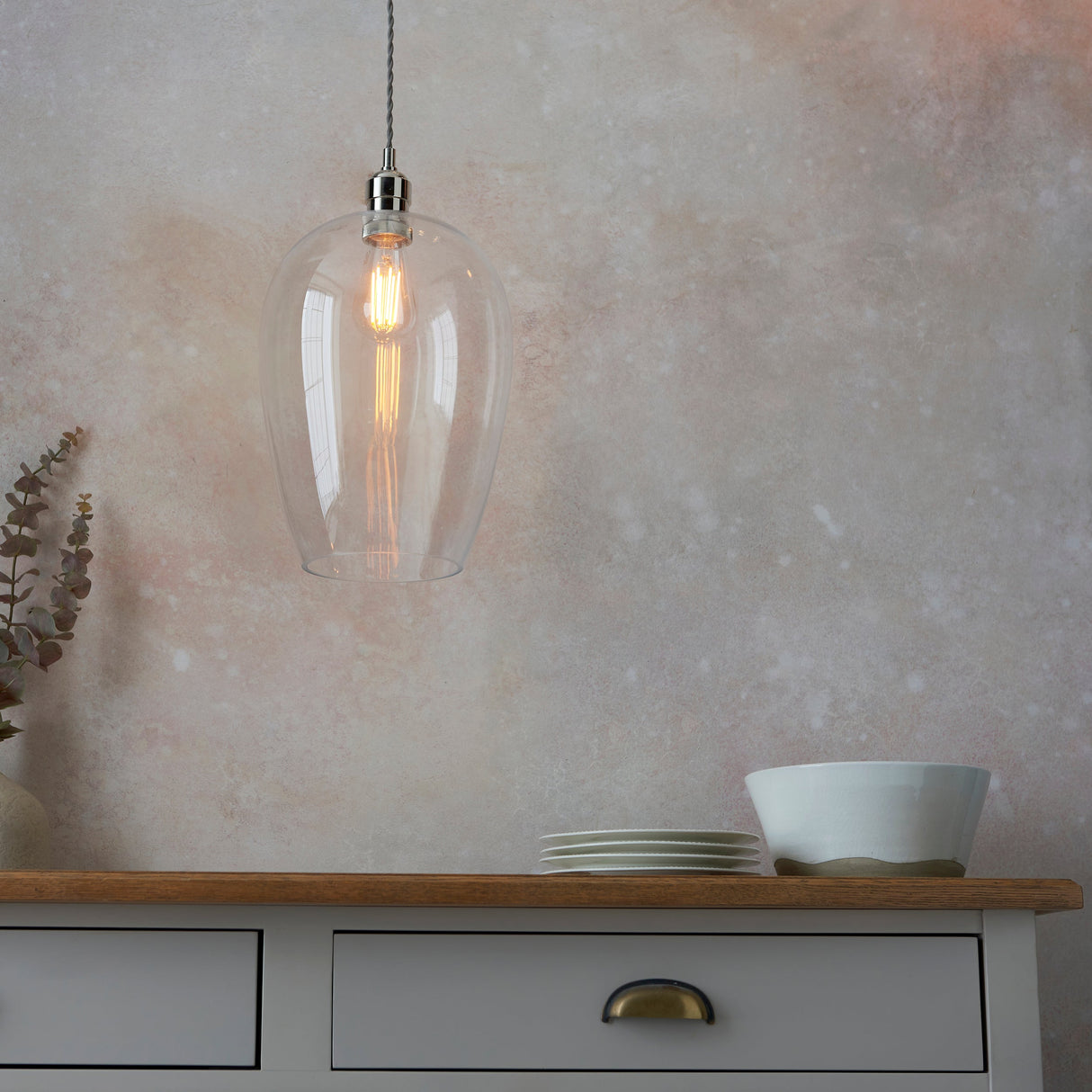 Vellmere Large Pendant in clear glass with bright nickel fitting and filament bulb, hung above a grey sideboard with plates and eucalyptus.