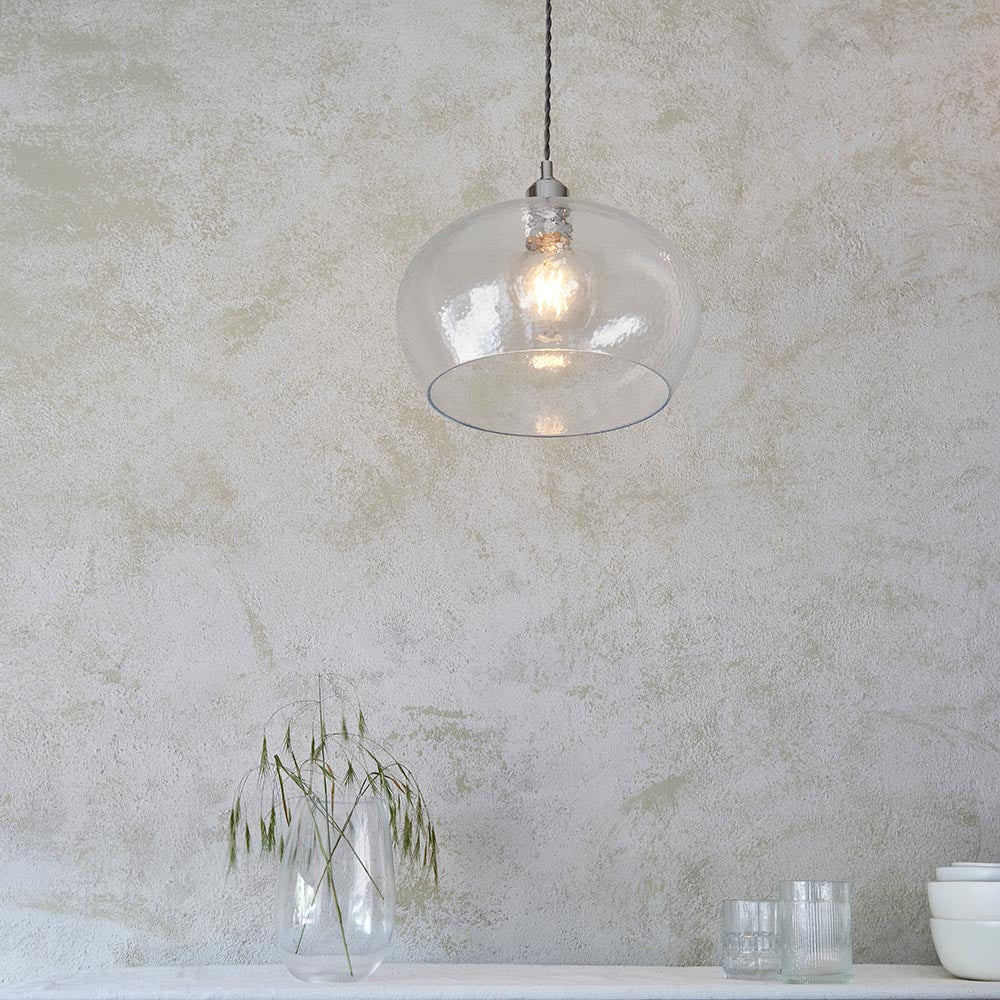 Draywick Hammered Medium Pendant in clear glass with bright nickel fitting from Lighting No.1, hung above a vase of grasses against a textured wall.