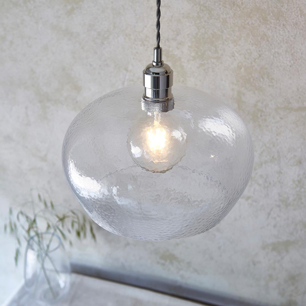 Draywick Hammered Medium Pendant from Lighting No.1 with a rounded clear hammered glass shade and bright nickel fitting, lit against a pale wall.