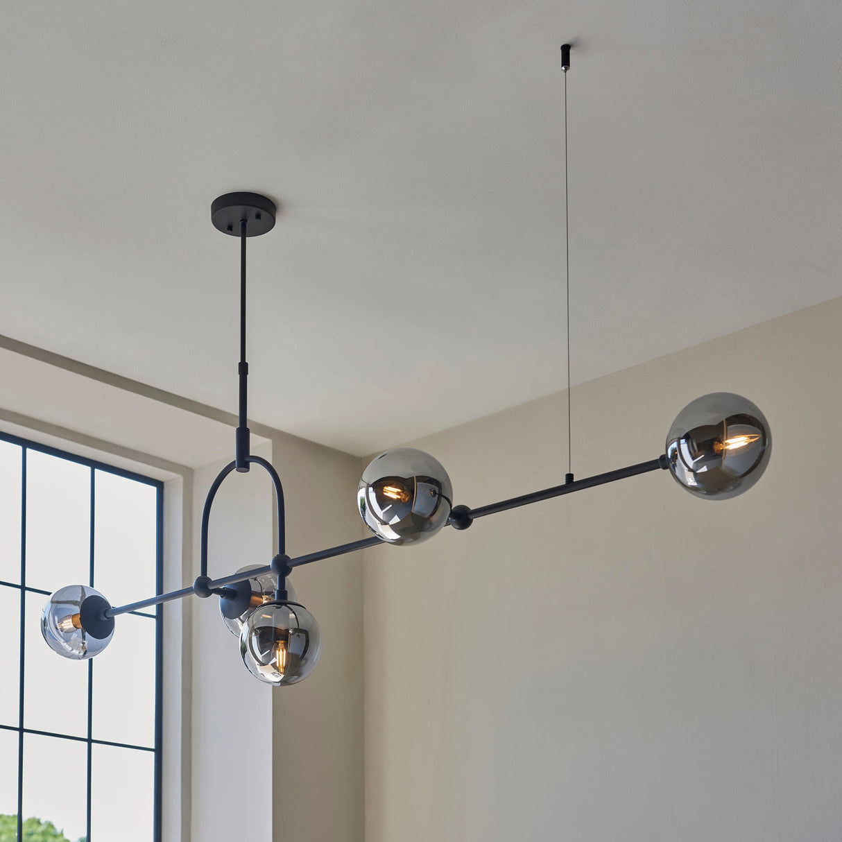 Haldmere linear pendant by Lighting No.1 with five smoked grey glass spheres along a matt black adjustable bar, shown in a bright room.