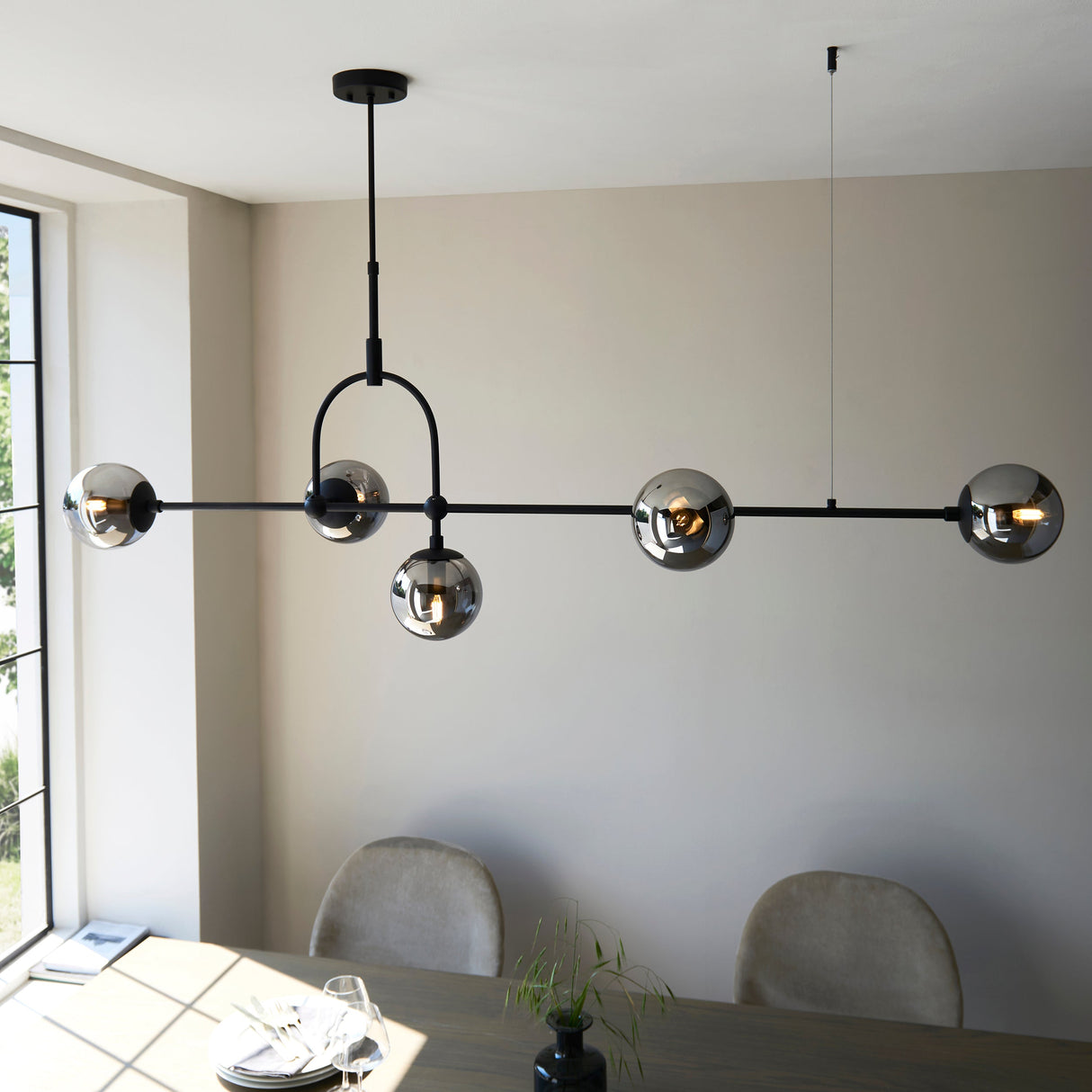 Lighting No.1 Haldmere linear pendant with five smoked grey glass spheres on a matt black bar and adjustable stem, above a dining table.