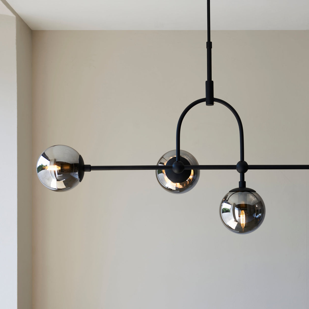 Haldmere pendant light by Lighting No.1 with three smoked grey glass spheres on a matt black bar and adjustable stem, shown in a neutral room.