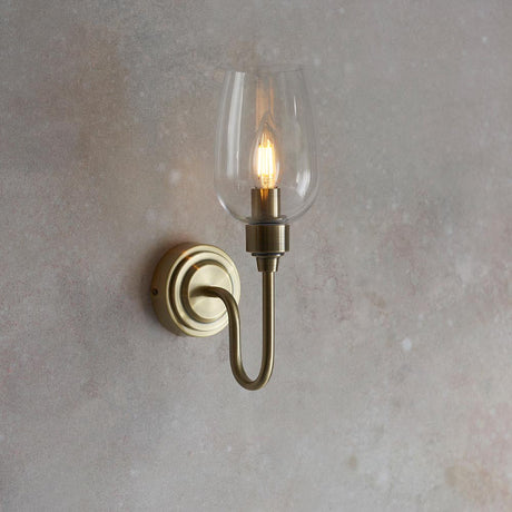Wraymoor single-light wall sconce from Lighting No.1 in antique brass with a slim clear glass shade and E14 candle bulb, mounted on a light grey wall.