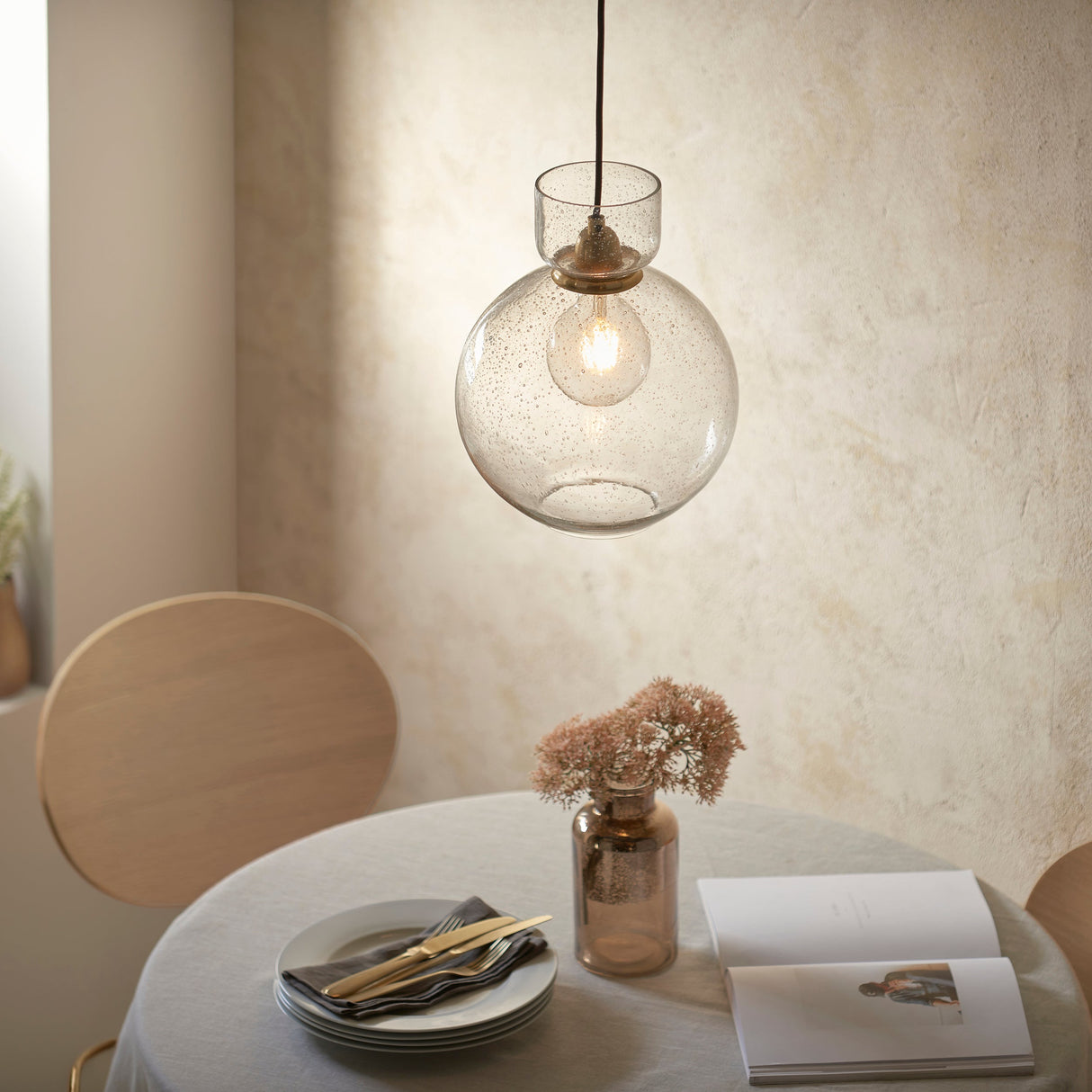 Yarnwell Bubble Pendant in clear bubbled glass with brass fitting by Lighting No.1, hung above a dining table set with plates, cutlery and a vase.