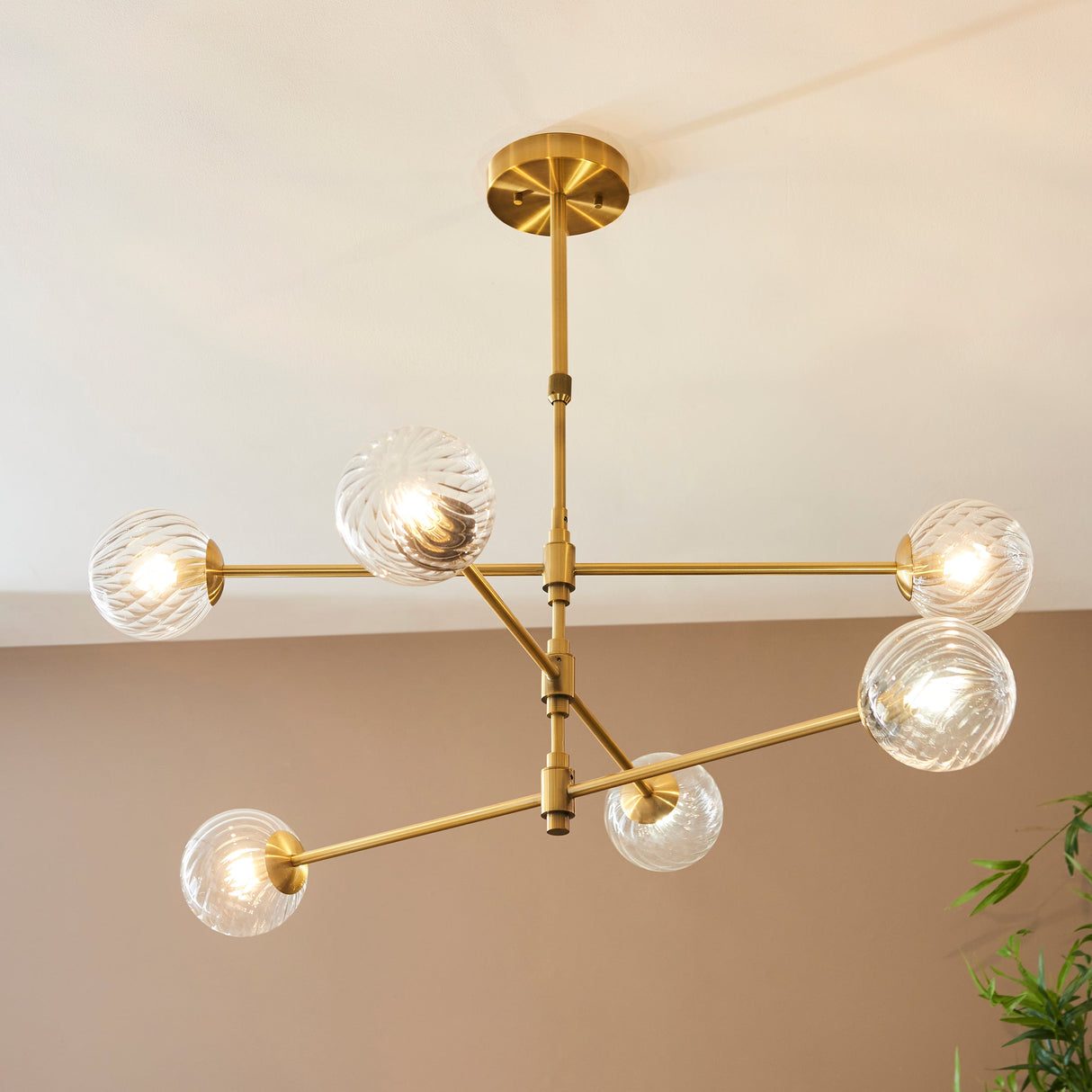 Mossgrove Twisted 6 Light Pendant in brushed aged brass with six angled arms holding twisted clear glass globe shades, mounted to a neutral ceiling.