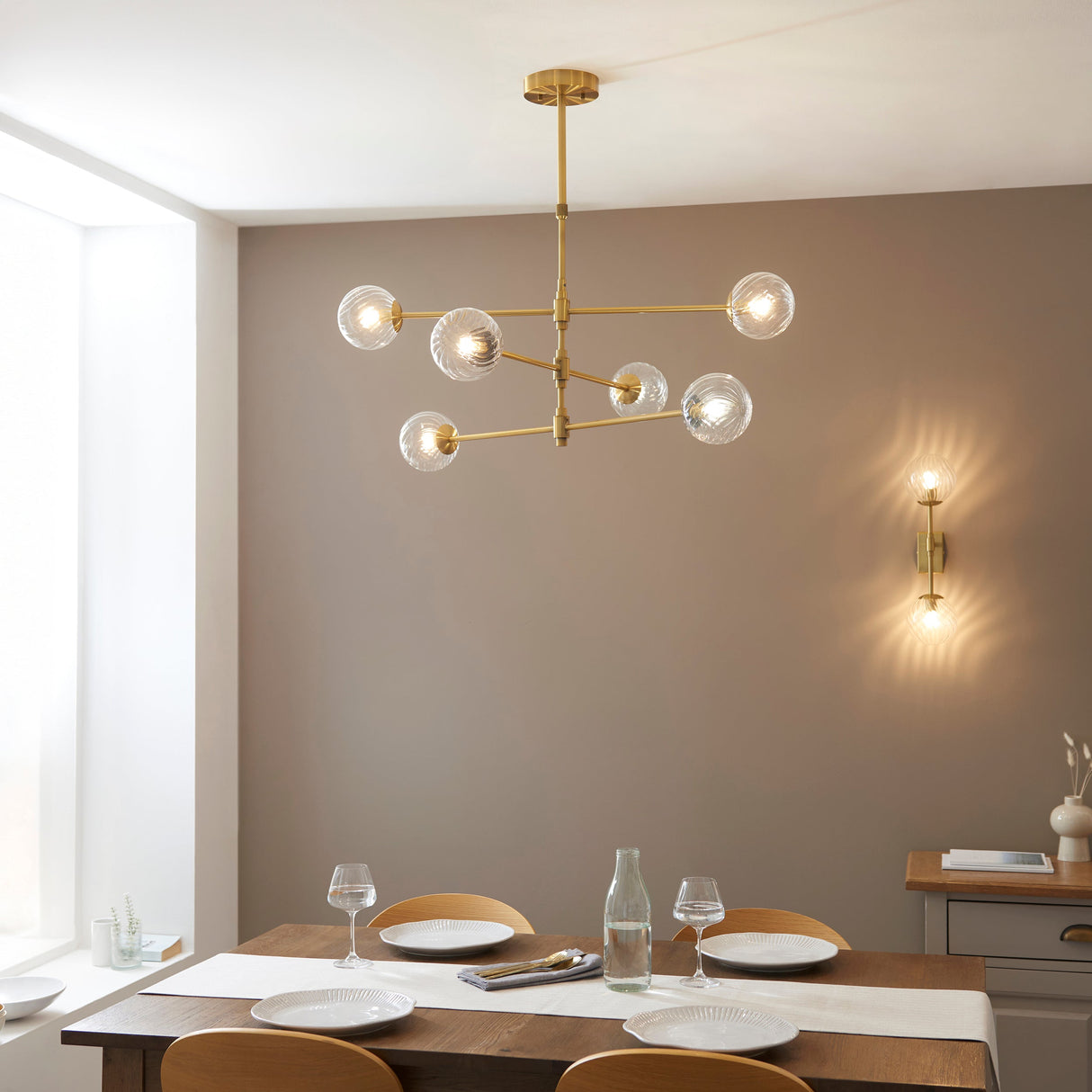 Mossgrove Twisted 6 Light Pendant in brushed aged brass with clear twisted glass globes, hung above a dining table beside a matching wall light.