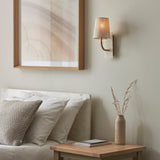 Fernhaven curved arm wall light in vintage brass with white marble backplate and tapered linen shade, mounted beside framed art above a bedroom headboard.