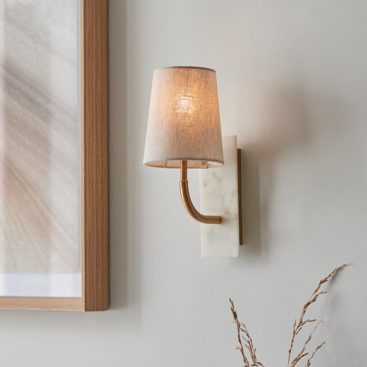Fernhaven curved arm wall light by Lighting No.1 with vintage brass arm, white marble backplate and tapered linen shade, mounted beside a wooden frame.