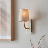 Fernhaven curved arm wall light by Lighting No.1 with vintage brass arm, white marble backplate and tapered linen shade, mounted beside a wooden frame.