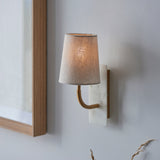 Fernhaven curved brass arm wall light with tapered linen shade on white marble backplate, mounted beside a wood-framed mirror on a pale grey wall.