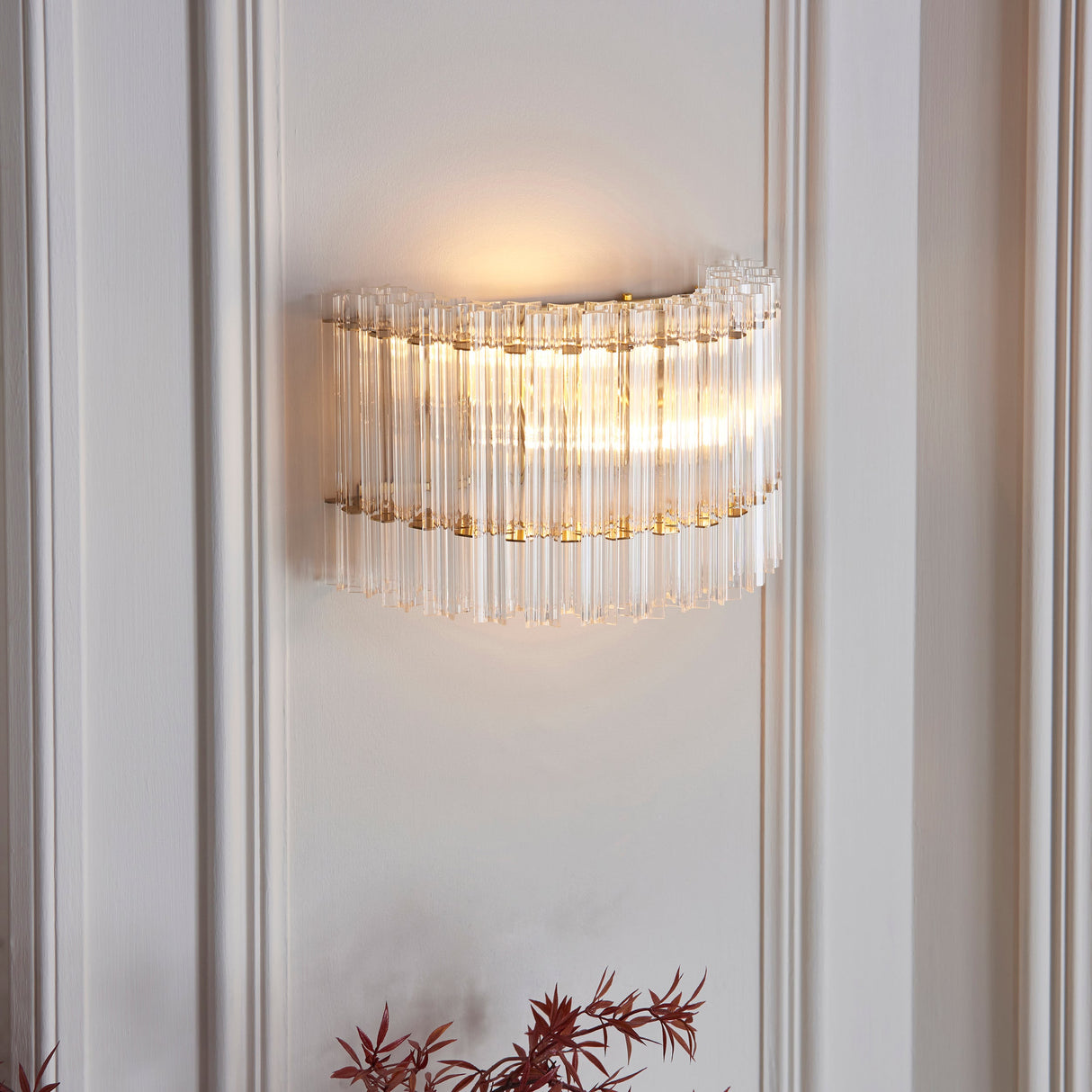 Lighting No.1 Nettlecote 2 light wall light illuminated on a panelled wall, with clear glass rods in a curved brushed aged brass frame.