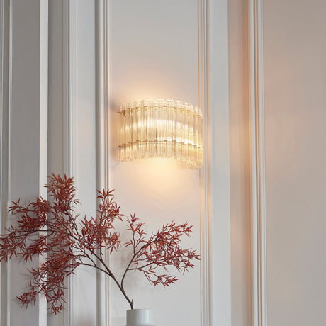 Nettlecote 2 light wall light in brushed aged brass with clear glass rods, illuminated on a panelled white wall above a vase of red foliage.