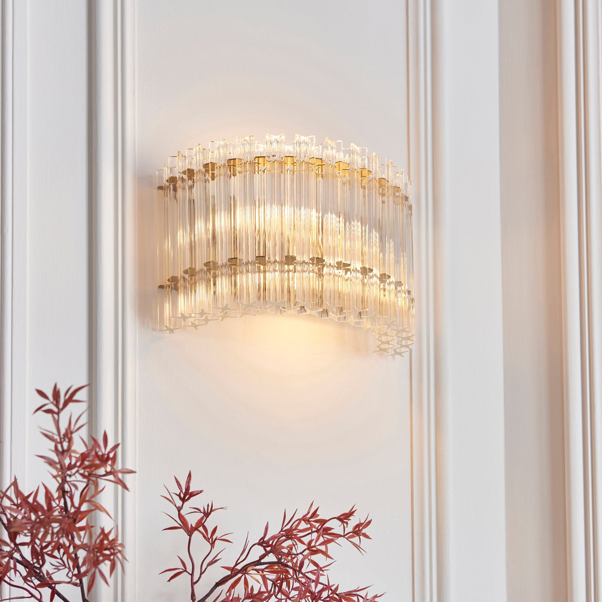 Nettlecote 2 light wall light by Lighting No.1, illuminated with clear glass rods in a brushed aged brass frame, mounted on a white panelled wall.