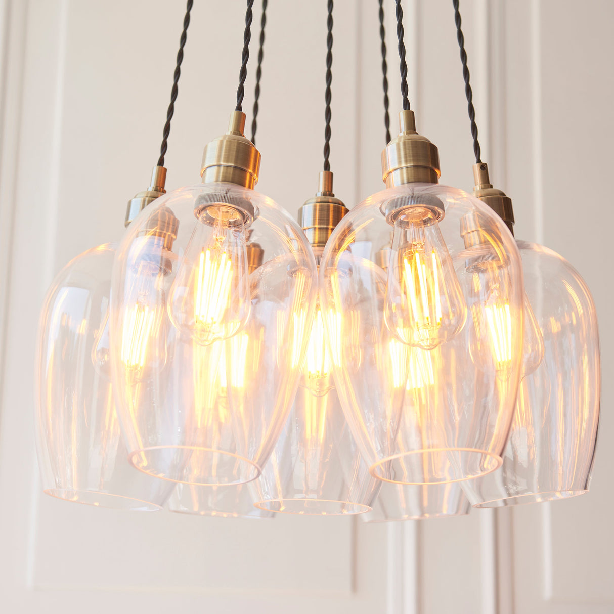 Quilbrook Cluster 7 Light Pendant from Lighting No.1 with clear glass globes and antique brass fittings, lit filament bulbs, on a pale panelled wall.