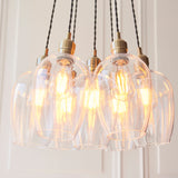 Quilbrook Cluster 7 Light Pendant from Lighting No.1 with clear glass globes and antique brass fittings, lit filament bulbs, on a pale panelled wall.