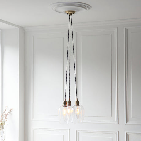 Lighting No.1 Quilbrook 3 light cluster pendant in antique brass with clear glass globes, hung from a ceiling rose against white panelled walls.
