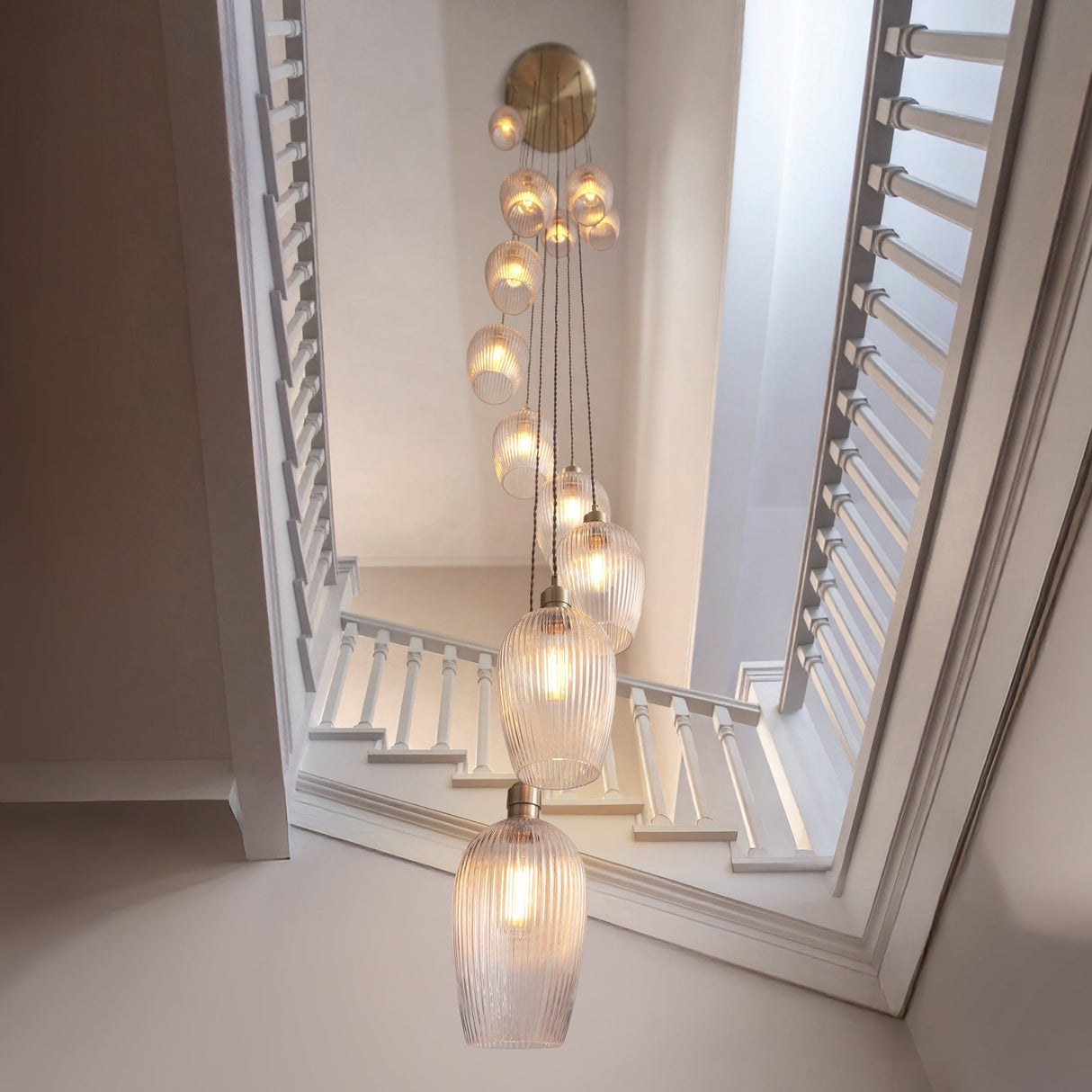 Fennhollow Ribbed Cascade 13 light pendant in antique brass with ribbed clear glass globes falling through a bright stairwell with white shutters.