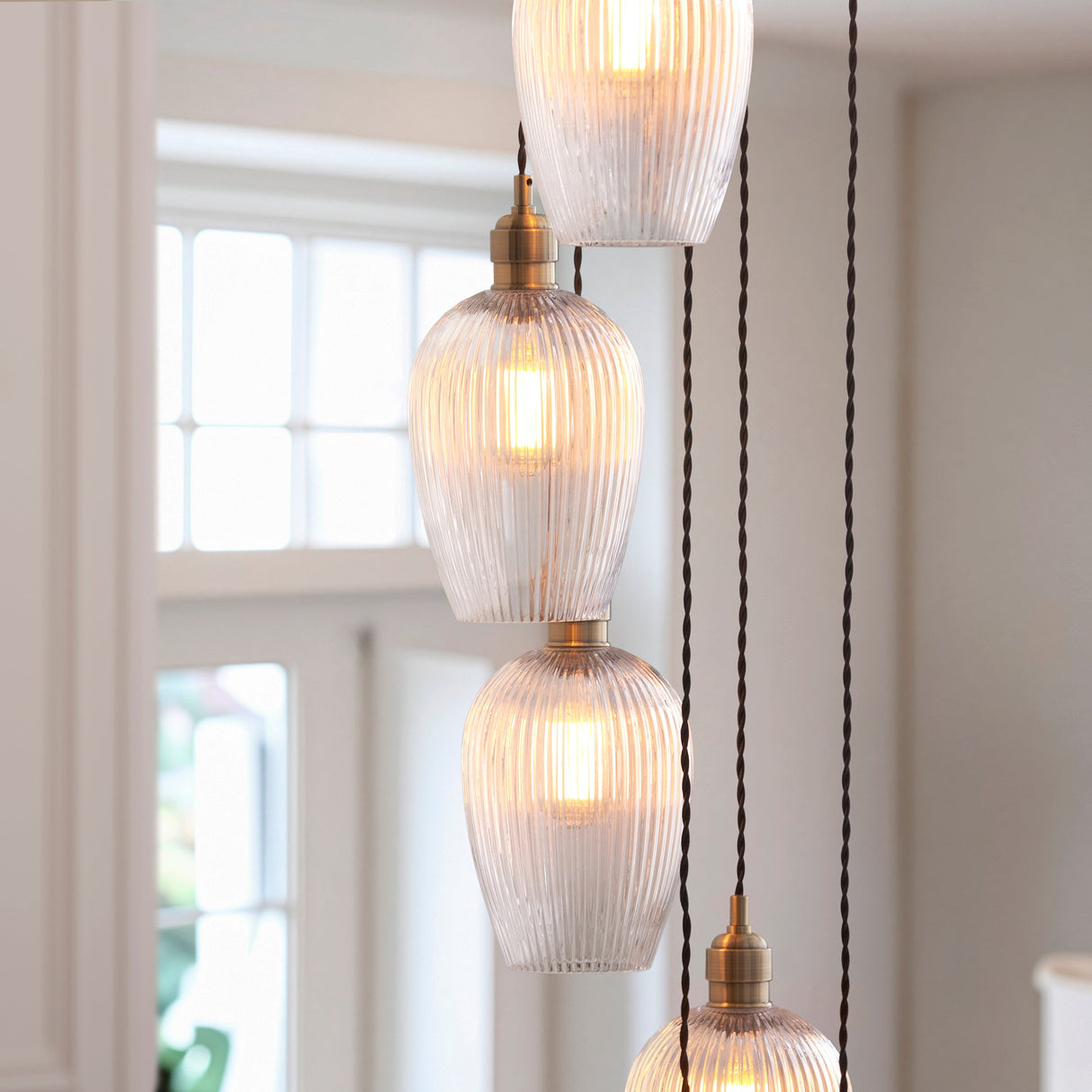 Fennhollow Ribbed Cascade 13 light pendant by Lighting No.1, with ribbed clear glass globes on antique brass fittings staggered near a bright window.
