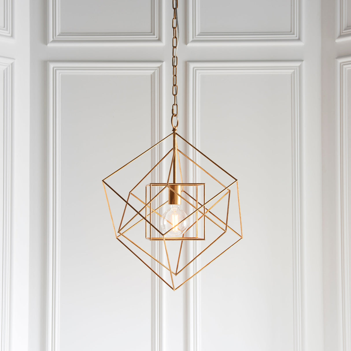 Padgrove Medium Angular Framed Single Pendant in gold leaf by Lighting No.1, hung on a chain against white panelled wall with exposed E27 bulb.