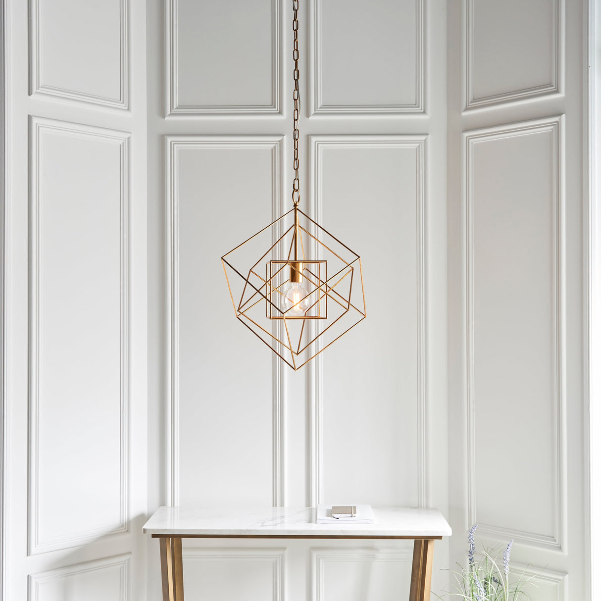Padgrove Medium Angular Framed Single Pendant in gold leaf by Lighting No.1, hung on a chain above a marble console against white panelled walls.