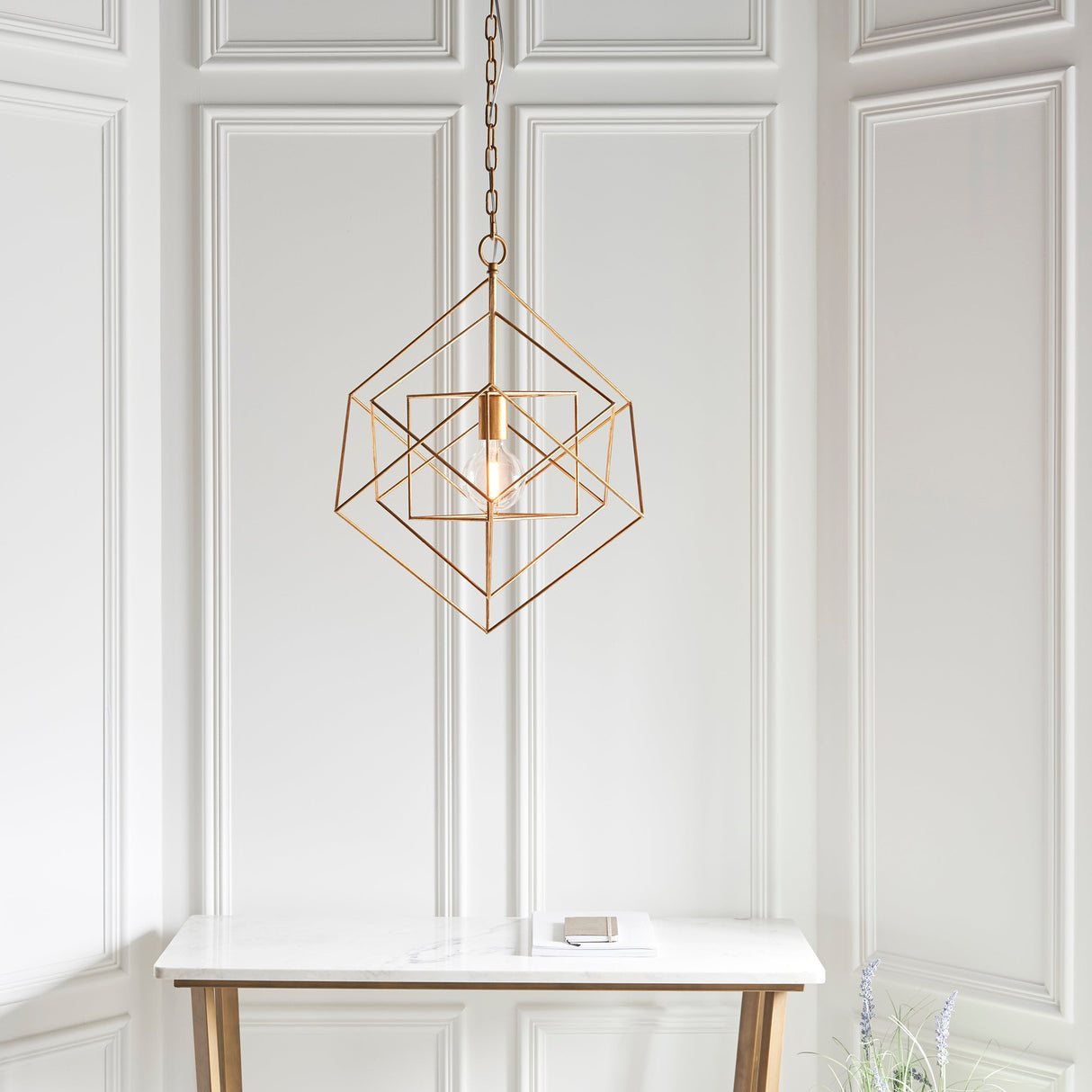 Padgrove medium angular framed single pendant in gold leaf by Lighting No.1, hung on a chain above a marble console in a panelled room.