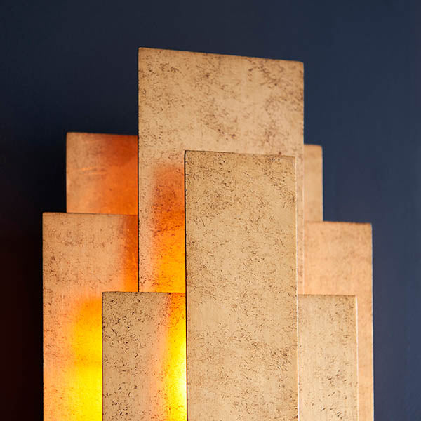 Dunmoor 2 light wall light from Lighting No.1 in gold, tiered rectangular metal panels with warm glow from concealed G9 lamps against a dark wall.