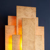 Dunmoor 2 light wall light from Lighting No.1 in gold, tiered rectangular metal panels with warm glow from concealed G9 lamps against a dark wall.