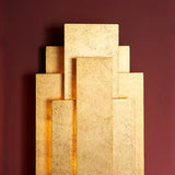 Dunmoor 2 light wall light from Lighting No.1 in gold, showing stacked rectangular metal panels in a tiered art deco layout against a deep red wall.