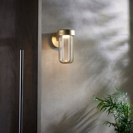 Yeldmoor brushed gold die-cast LED outdoor wall light with clear glass, mounted on a concrete wall beside a dark wood door with a leafy plant.