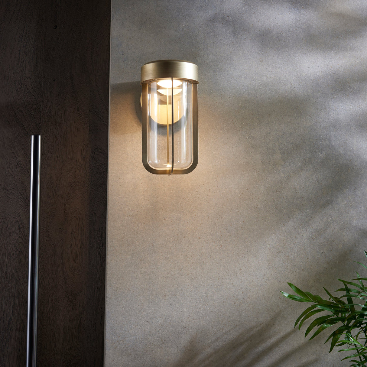 Yeldmoor brushed gold die-cast LED outdoor wall light with clear glass, mounted on a render wall beside dark wood cladding and a green plant.