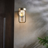 Yeldmoor brushed gold die-cast LED outdoor wall light with clear glass, mounted on a render wall beside dark wood cladding and a green plant.
