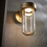 Yeldmoor die-cast LED outdoor wall light in brushed gold with clear glass cylinder shade, mounted on a rendered wall and casting warm up and down light.