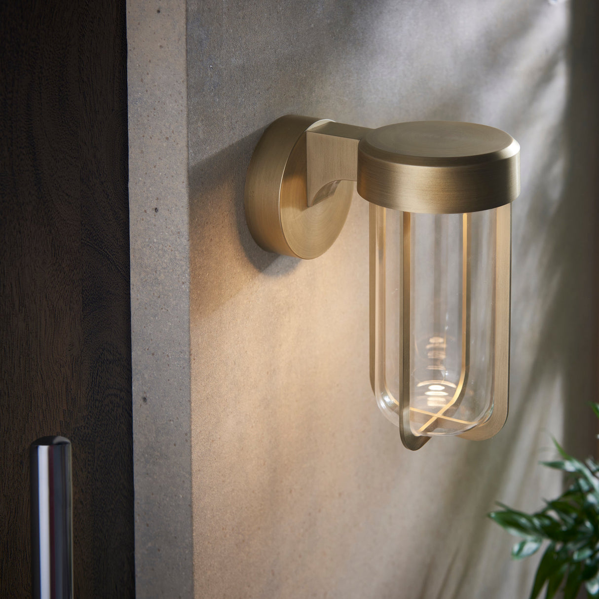Yeldmoor die-cast LED outdoor wall light in brushed gold with clear glass shade, mounted on a concrete wall beside a dark timber door.