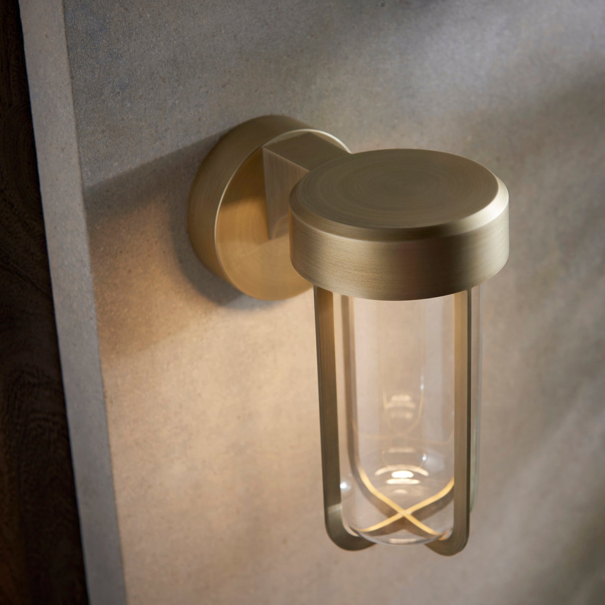 Yeldmoor brushed gold die-cast LED outdoor wall light by Lighting No.1, mounted on a stone wall with clear glass cylinder casting warm ambient light.