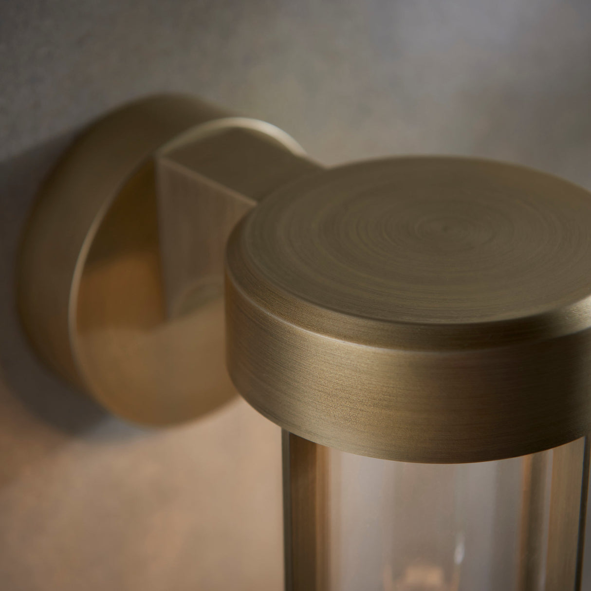 Close-up of Lighting No.1 Yeldmoor die-cast outdoor wall light showing brushed gold circular top, wall bracket, and clear glass cylinder.