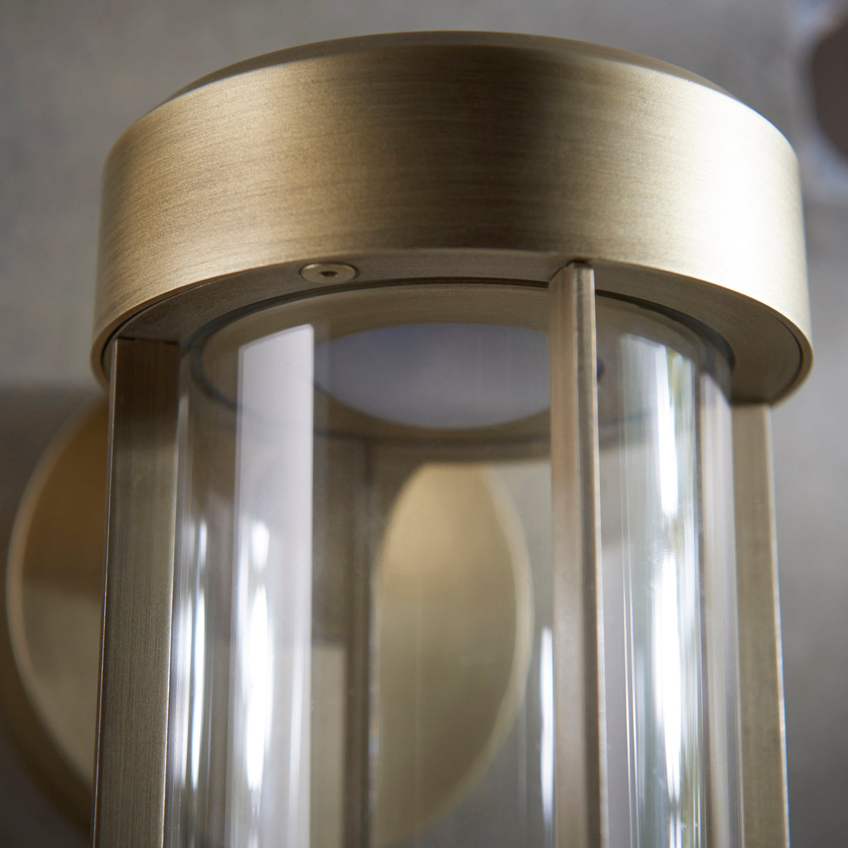 Close-up of Lighting No.1 Yeldmoor die-cast LED outdoor wall light showing brushed gold cylindrical top and clear glass cylinder shade.