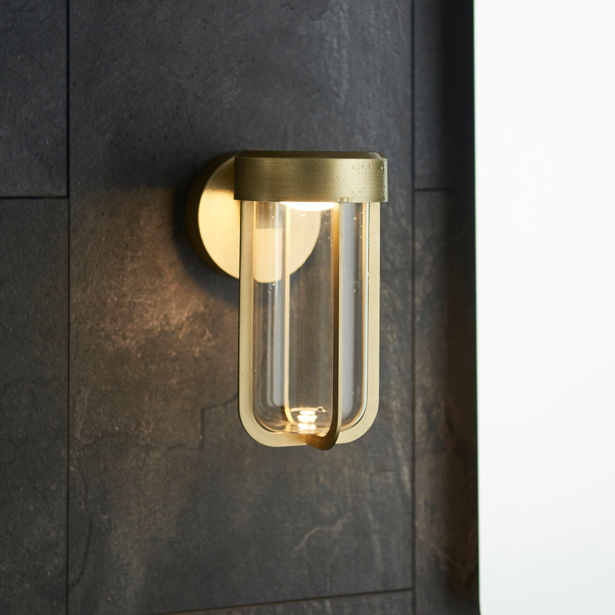 Yeldmoor brushed gold die-cast LED outdoor wall light with clear glass tube, mounted on a dark slate wall beside a doorway.