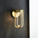 Yeldmoor brushed gold die-cast LED outdoor wall light with clear glass tube, mounted on a dark slate wall beside a doorway.