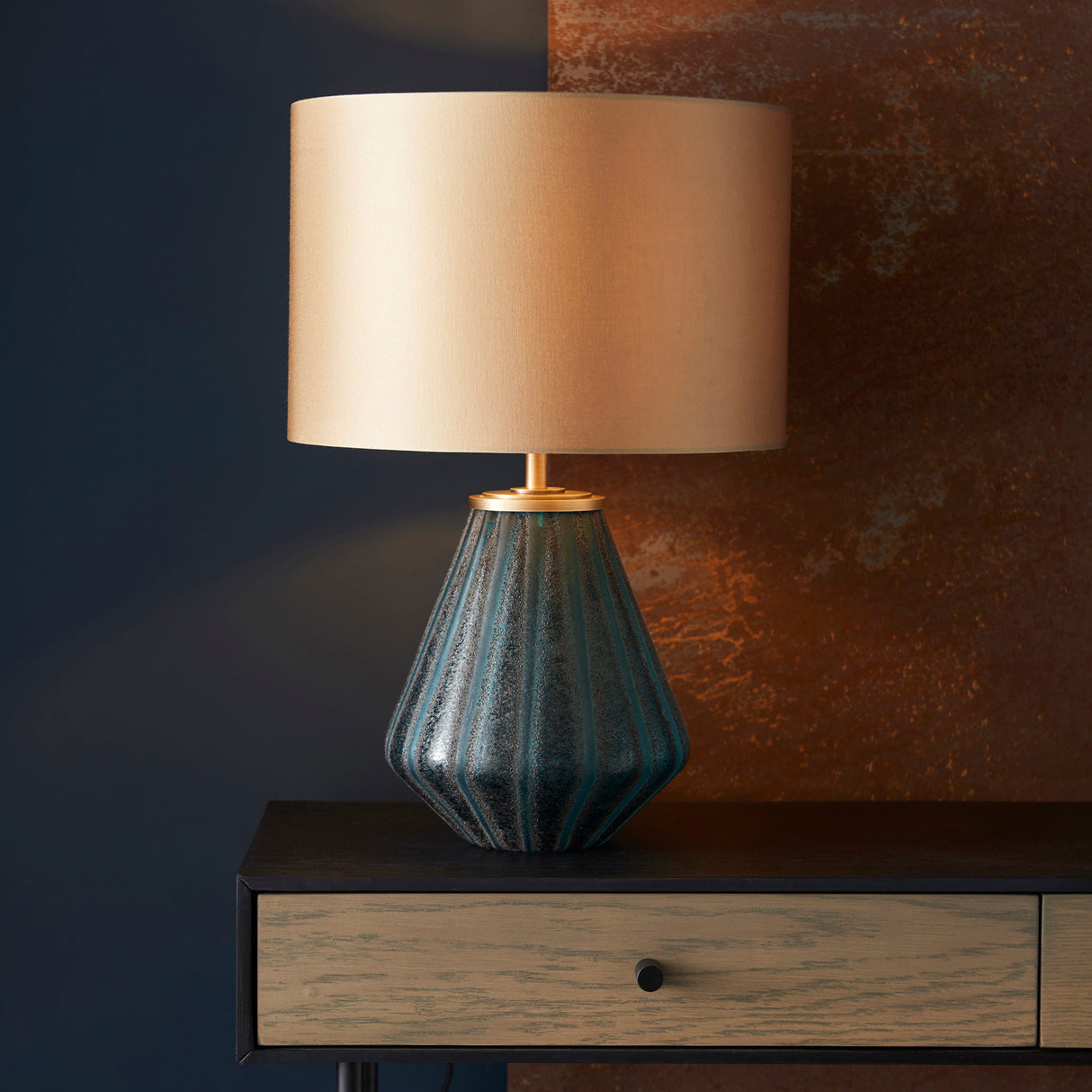 Foxleigh Table Lamp by Lighting No.1 with faceted turquoise glass base and cream drum shade on a dark console against a navy and rust wall.