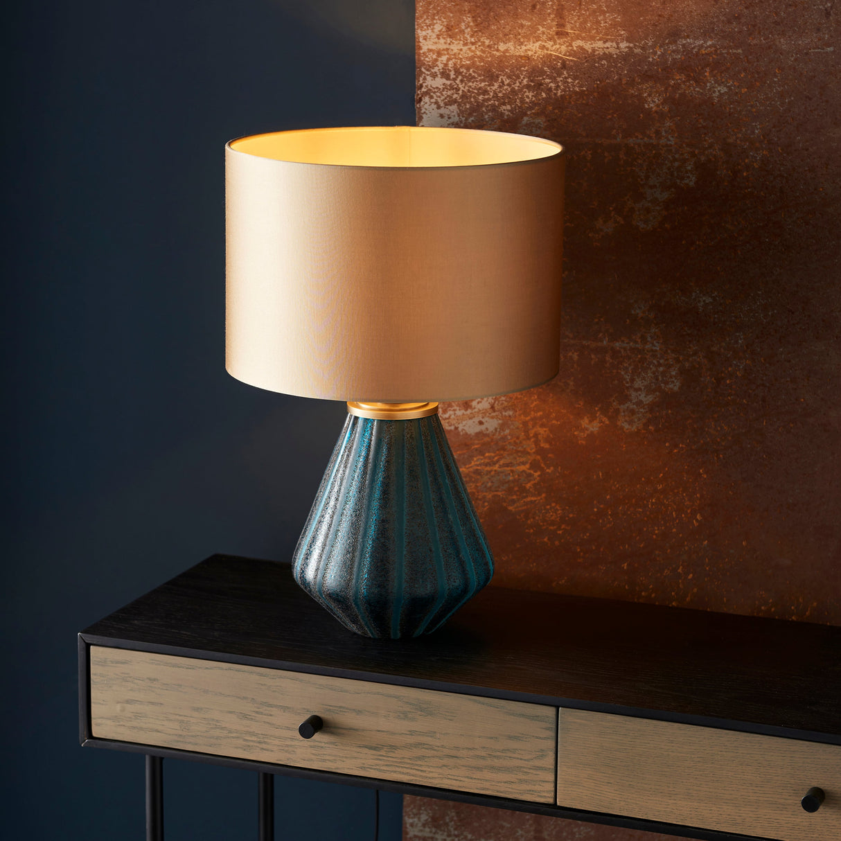 Foxleigh table lamp by Lighting No.1 with turquoise faceted glass base and gold drum shade, on a dark wood console against a navy and rust wall.