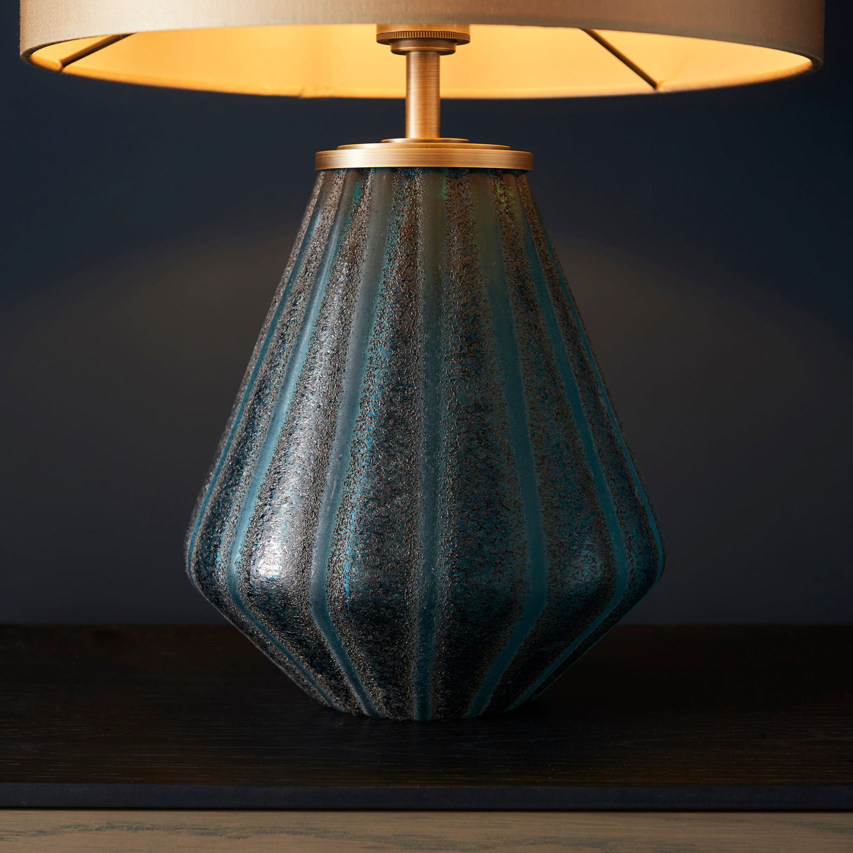Foxleigh table lamp from Lighting No.1 with a ribbed turquoise glass base and brushed gold fittings beneath a lit gold shade on dark wood.