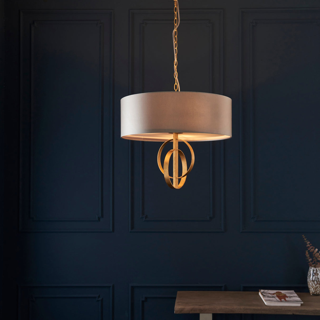 Sparrowmere 3 light pendant by Lighting No.1 with mink fabric drum shade and gold leaf double hoops, hung from a chain against a dark blue panelled wall.