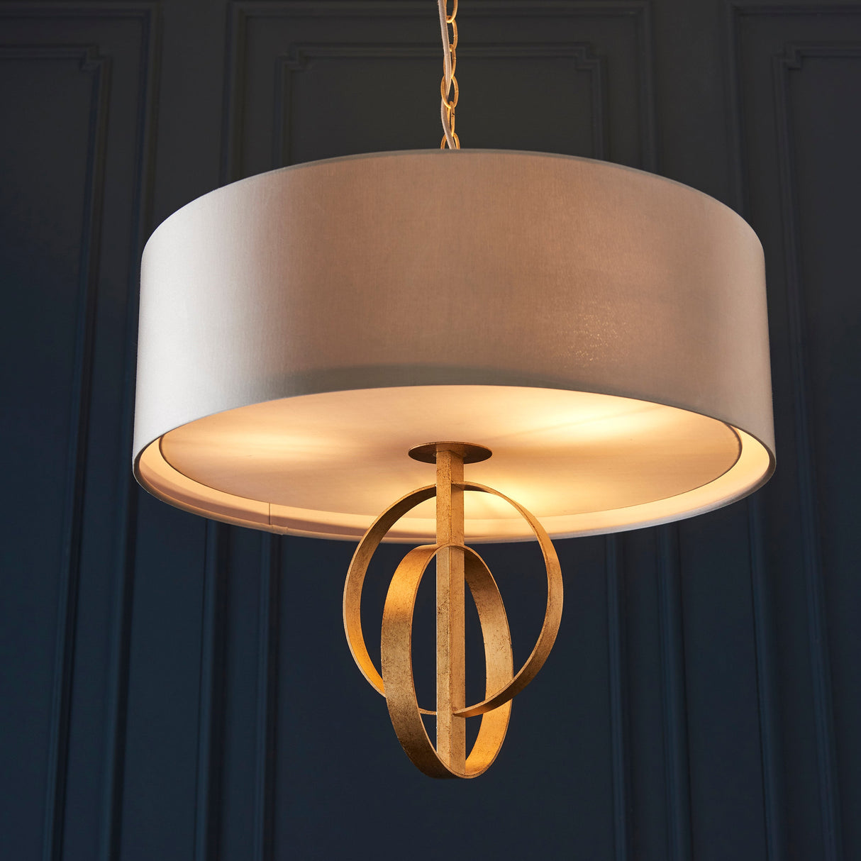 Sparrowmere Double Hoop 3 Light Pendant by Lighting No.1 with mink fabric drum shade and gold leaf hoops, suspended against a dark blue panelled wall.