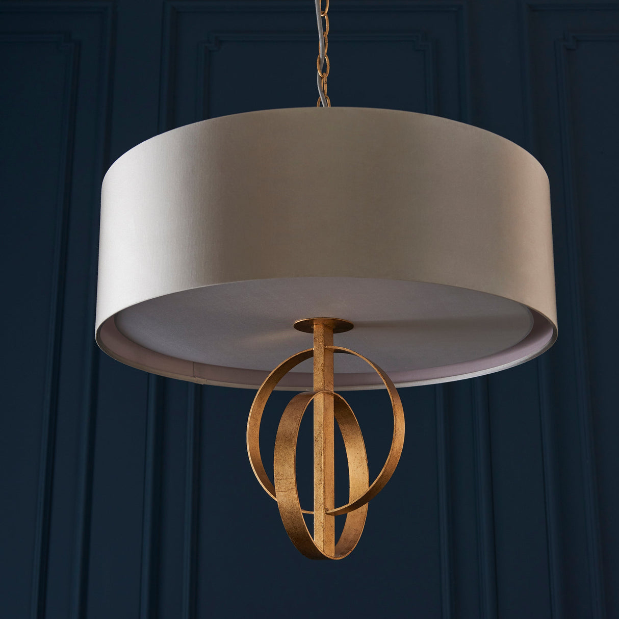 Sparrowmere Double Hoop 3 light pendant from Lighting No.1, mink fabric drum shade with gold leaf metal hoops, shown against a dark blue panelled wall.