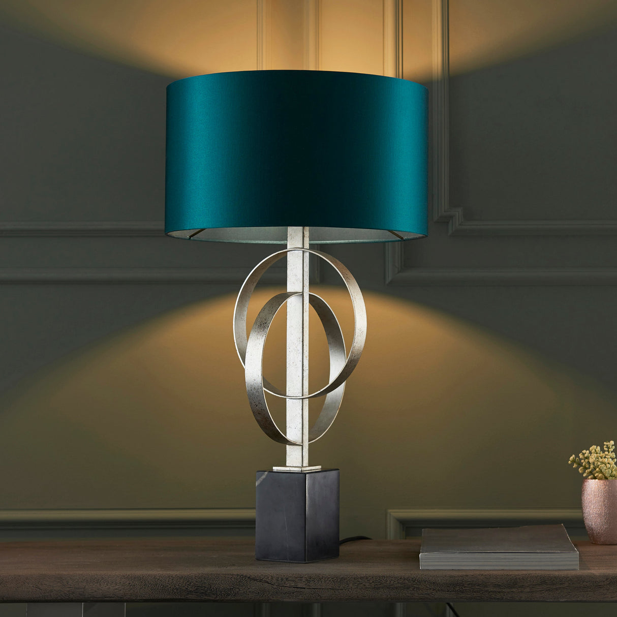Sparrowmere Double Hoop table lamp with silver leaf interlocking hoops, teal fabric drum shade and dark square base on a wooden console.